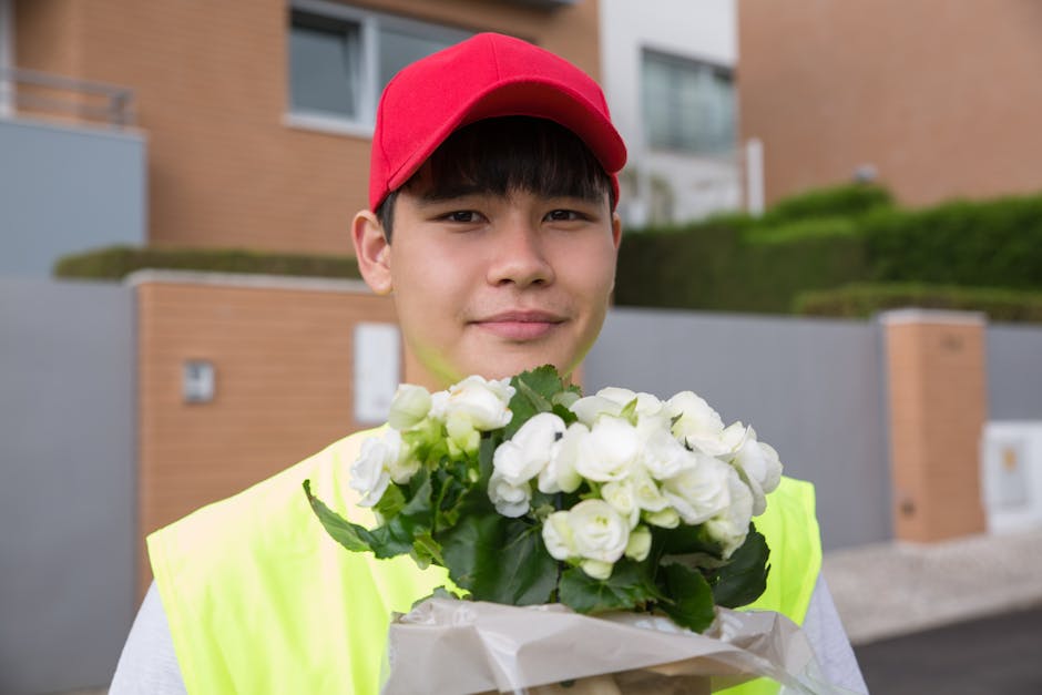 A young delivery person wearing a bright yellow high-visibility vest and a red cap is standing outdoors in front of modern residential buildings, holding a bouquet of fresh white roses wrapped in whit