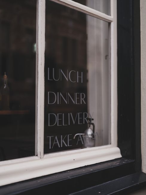A close-up view of a classic white-framed window with clear glass panes, displaying white text listing meal options such as 'Lunch,' 'Dinner,' 'Delivery,' and 'Takeaway' on the interior side. Behind t