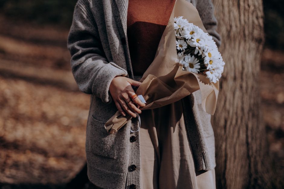 A person dressed in a grey cardigan and beige pants is holding a bouquet of fresh white daisies wrapped in light brown paper, with a glimpse of a dark top underneath. The bouquet is arranged with sign