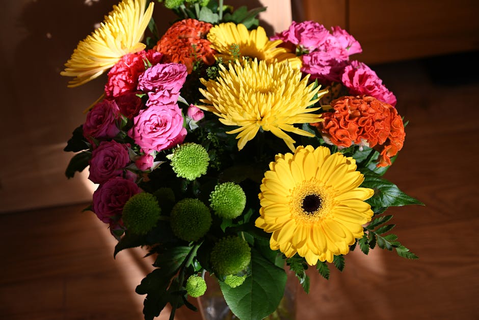 A vibrant bouquet of fresh flowers arranged in a glass vase on a wooden surface, featuring large yellow gerbera daisies, bright orange carnations, pink lisianthus, and round green chrysanthemums, with