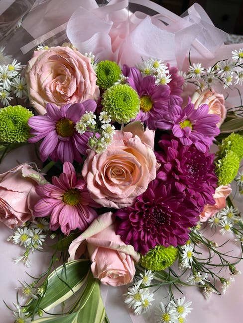 A close-up view of a floral bouquet featuring light pink roses with soft, layered petals, vibrant purple daisies with yellow centers, bright green button chrysanthemums, and small white daisy-like flo