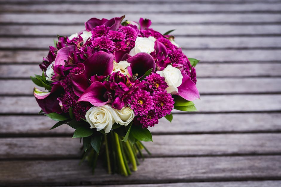 A round bouquet of fresh flowers arranged with pink calla lilies, white roses, and vibrant purple chrysanthemums. The bouquet features lush green foliage interspersed among the blooms, wrapped with el